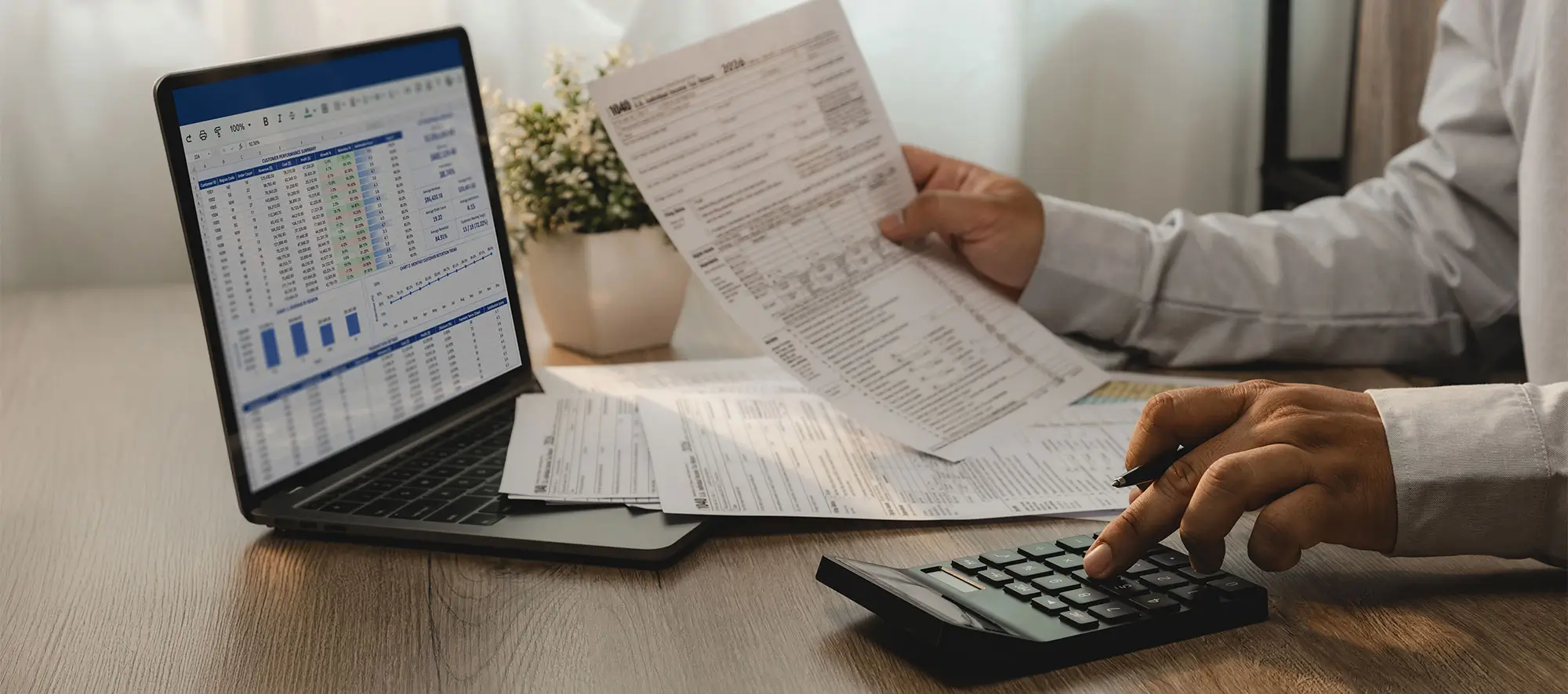 Accountant reviewing financial report and tax documents on laptop while using calculator for account analysis.