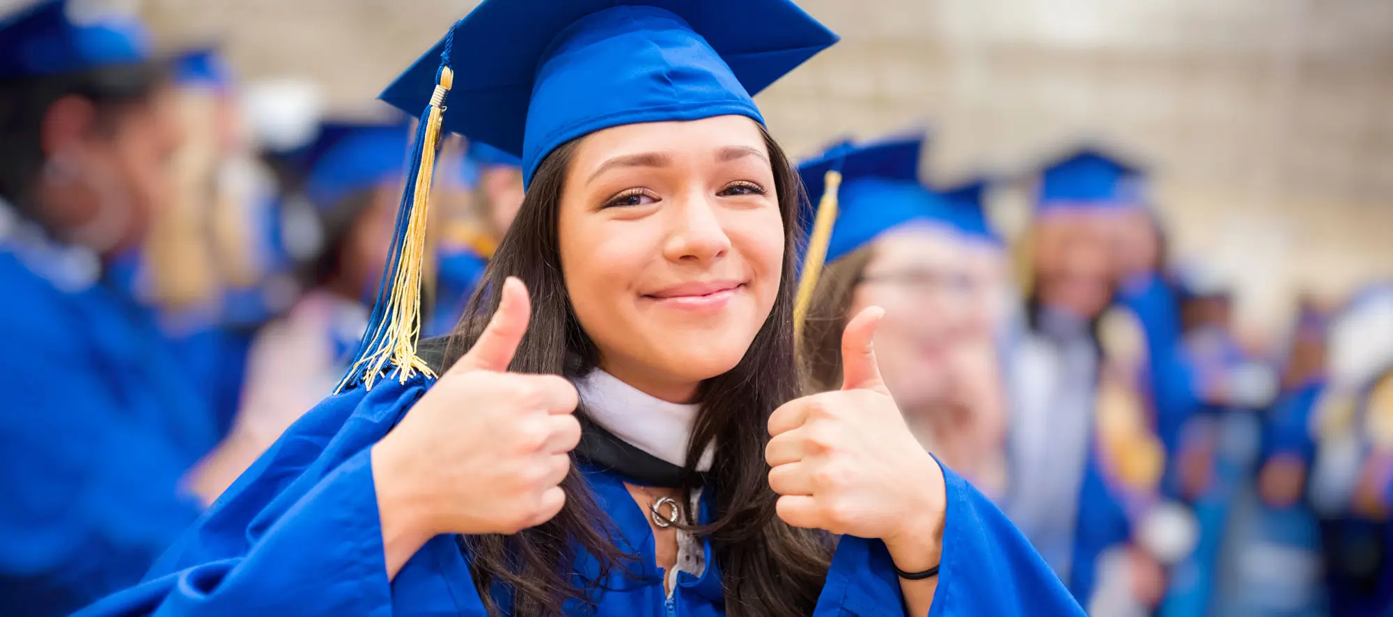 Young college girl in cap and gown at the graduation ceremony giving the thumbs up for success.