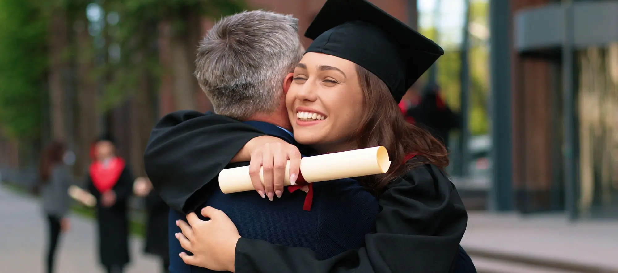 Happy female graduate are embracing with her father with diploma at her hands and rejoicing with each other. Man is hugging his daughter with other students moving and talking in background